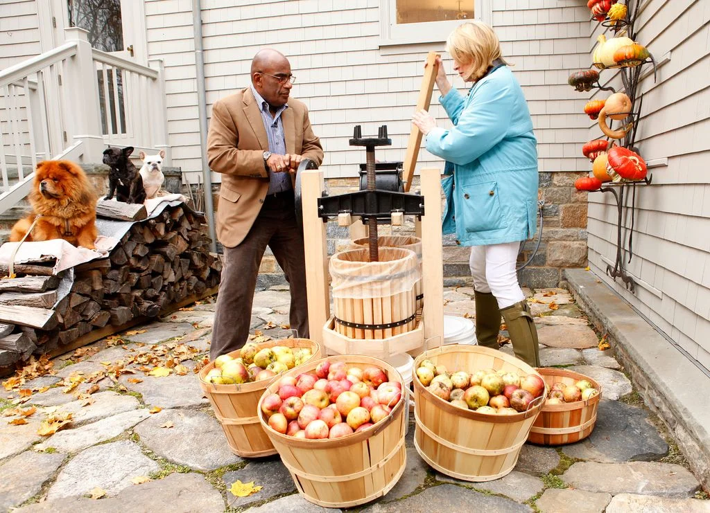 Martha Stewart pressing apples right beside Al Roker standing on stone paved area four large baskets of apples in...