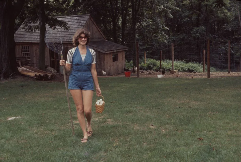 Martha Stewart holding basket of eggs and chicken wire net attached to pole as she walks across lawn wood ADU in background
