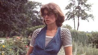Martha Stewart in denim overalls and striped shirt holding basket filled with eggs standing on gravel path through...