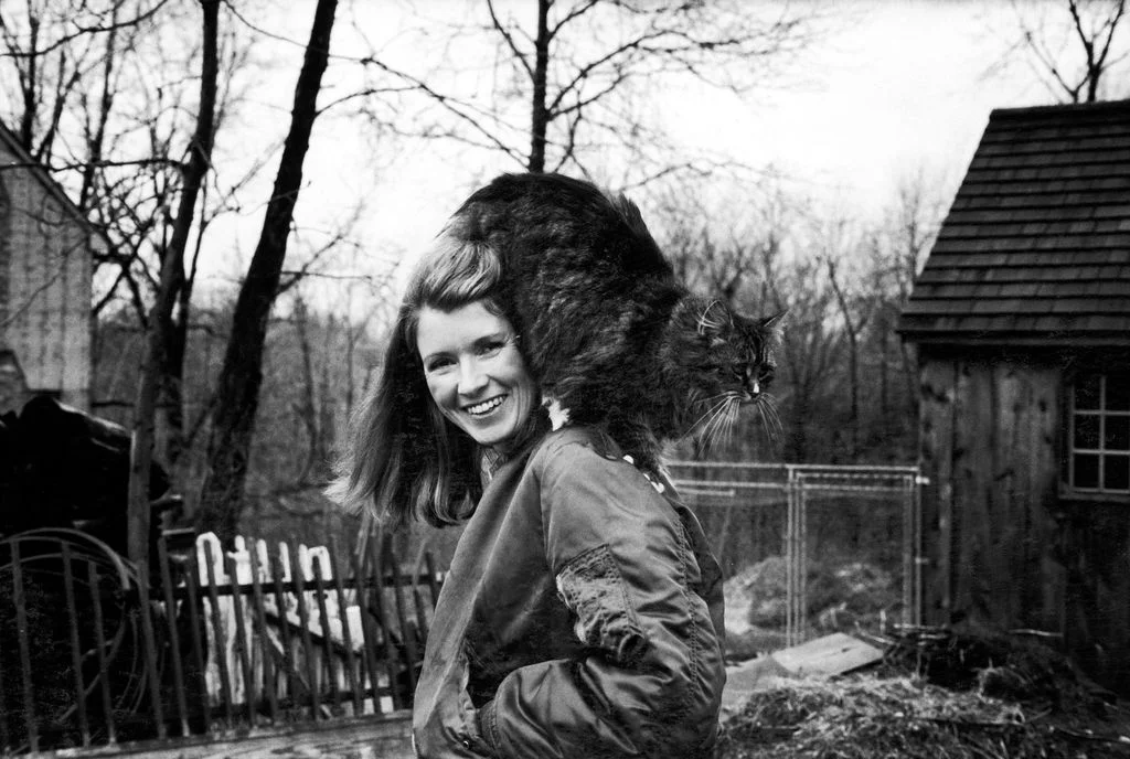 blackandwhite photo of Martha Stewart smiling with a cat perched on her shoulder while standing outside on the grounds...