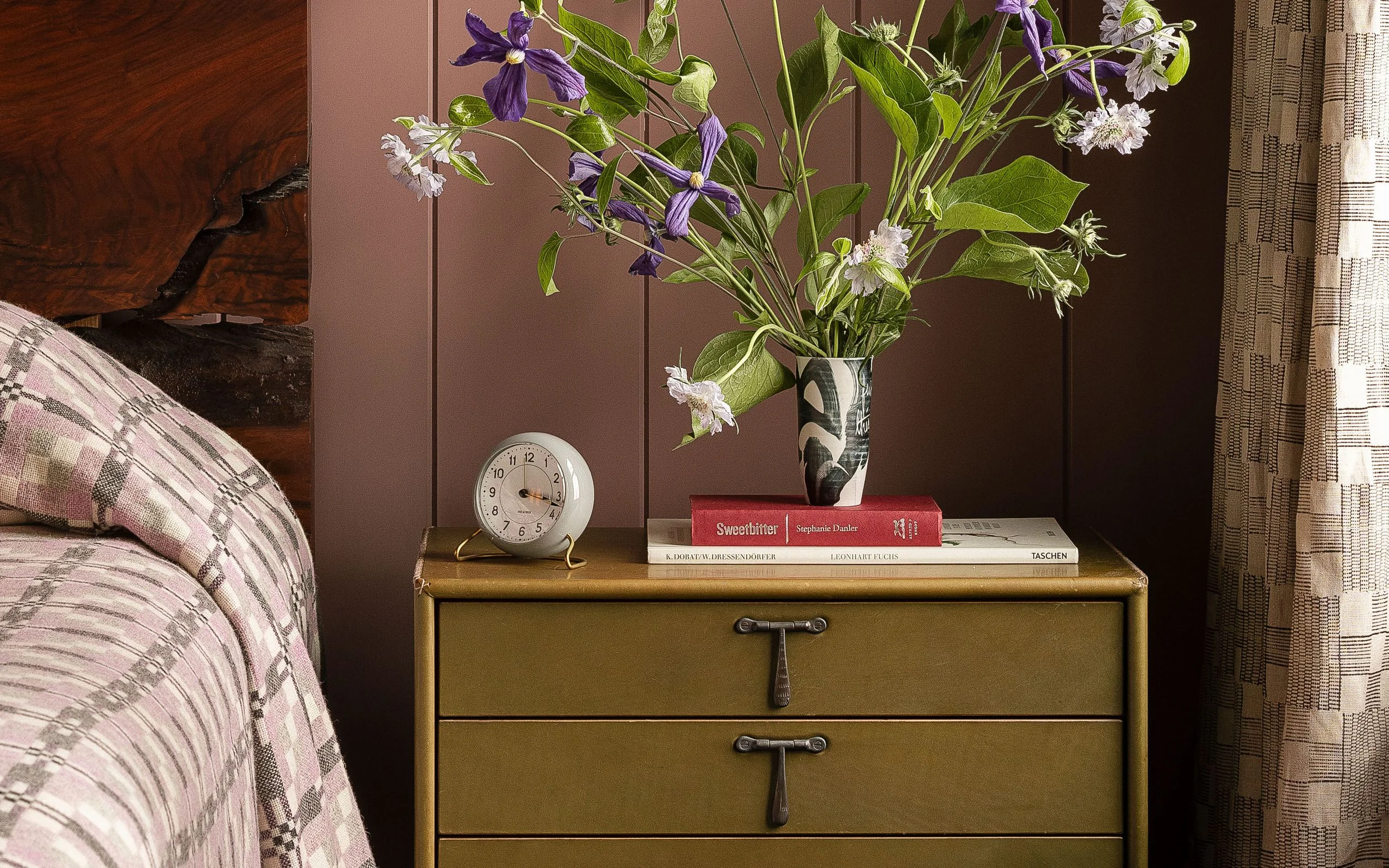 Bedside table with an alarm clock and vase of flowers on it.