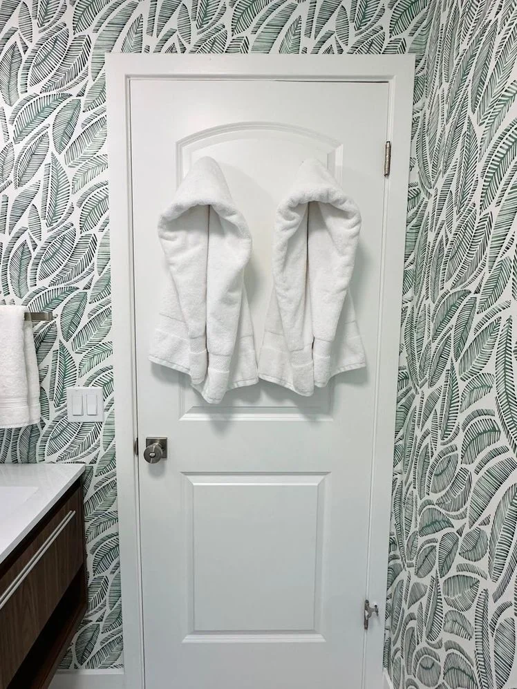 Two white towels hanging on the back of a door inside a bathroom with tropical leaf wallpaper.