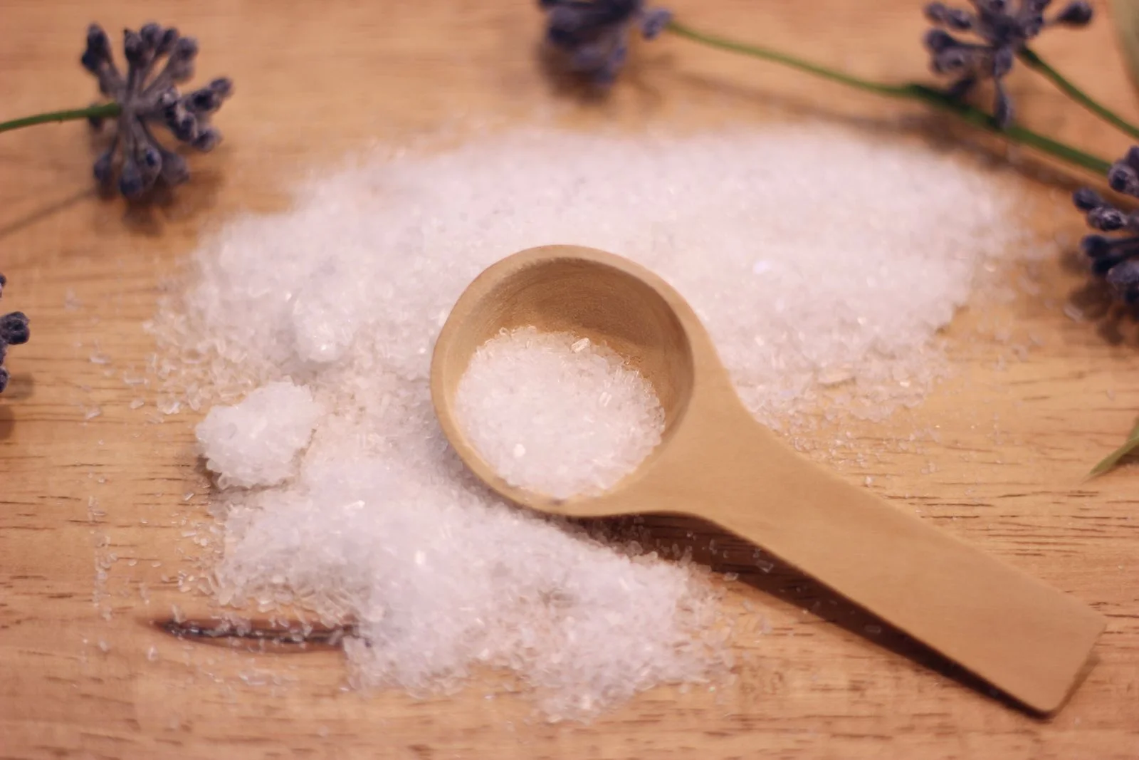 small wood spoon full of epsom salt lying in pile of epsom salt spilled onto wood surface purple flowers