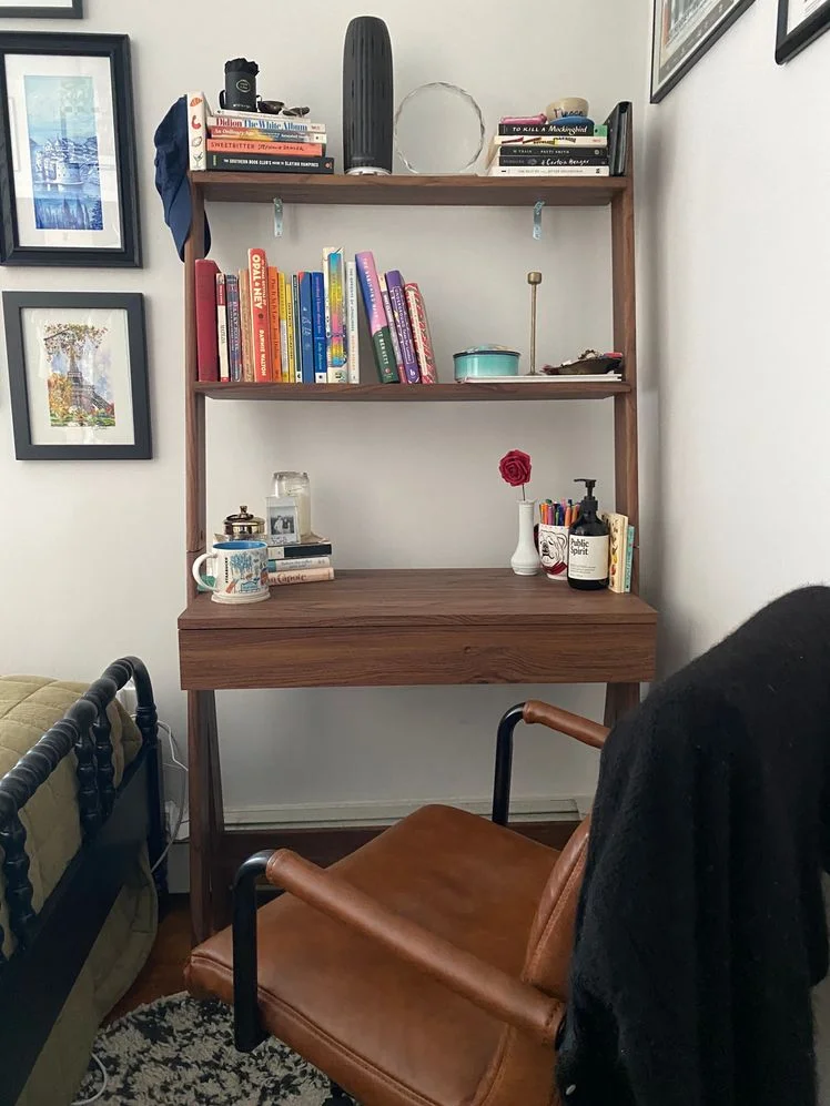 Wooden desk with a 2-tier ladder shelf attached to the top styled with books and art with a desk chair in front of it.
