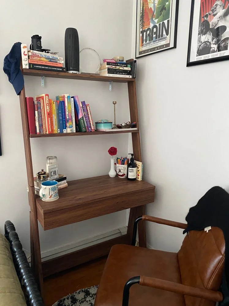 Wooden desk with a 2-tier ladder shelf attached to the top styled with books and art. A leather desk chair sits in front of it.