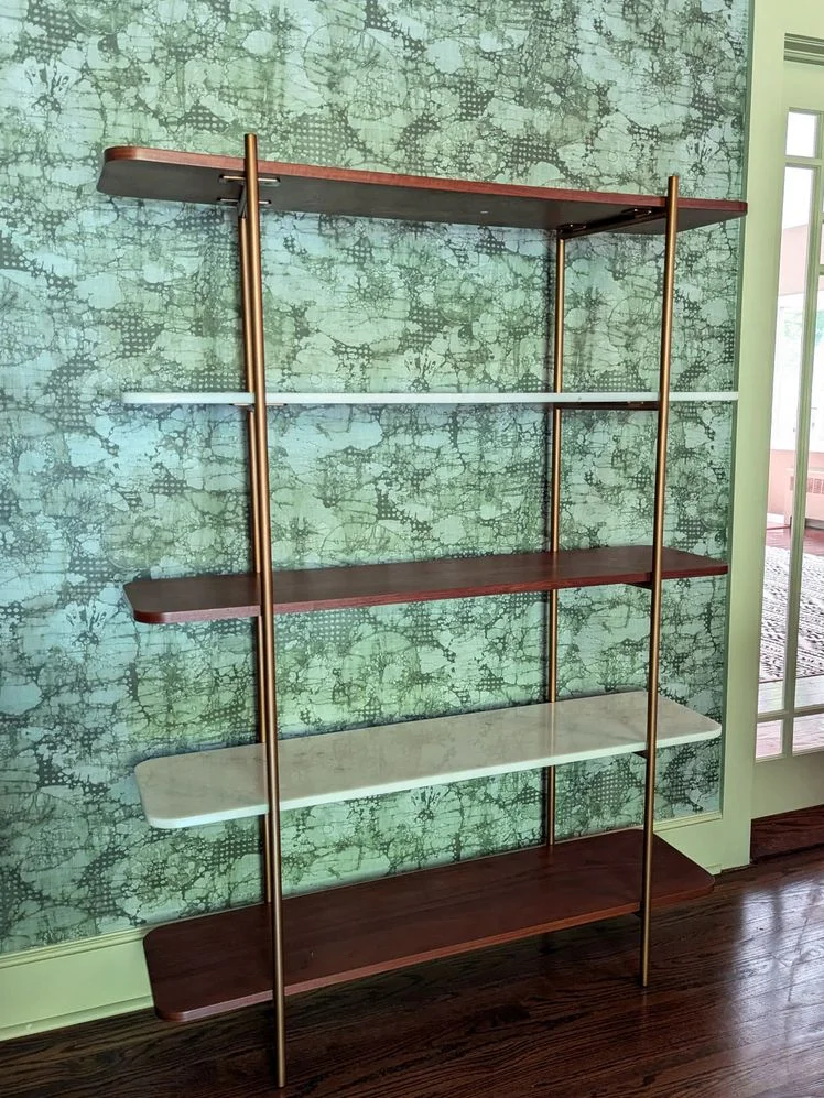 5-tier bookshelf with alternating wood and marble shelves and a gold metal frame in front a green wallpapered wall.