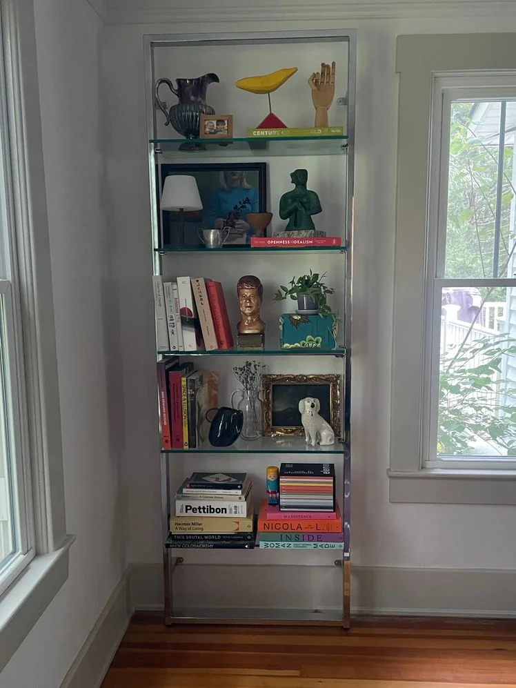 Wall mounted bookshelf with a chrome frame and glass shelves in the corner of a room between two windows, styled with various books, sculptures, and art.