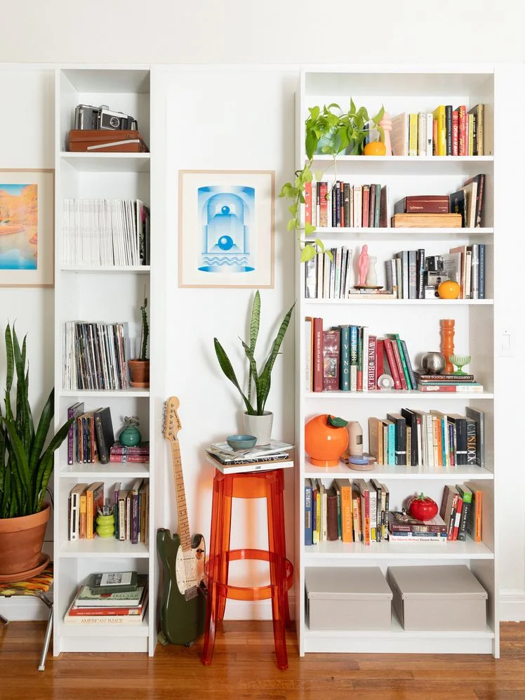Two white IKEA bookshelves in a living room, one narrow and one wide, styled with various books and knick knacks.