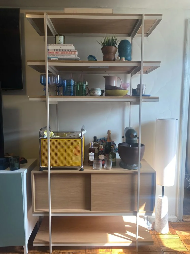 Wood and metal shelving unit with 5 tiers and one sliding door cabinet holding various kitchen and decor items in a living room.