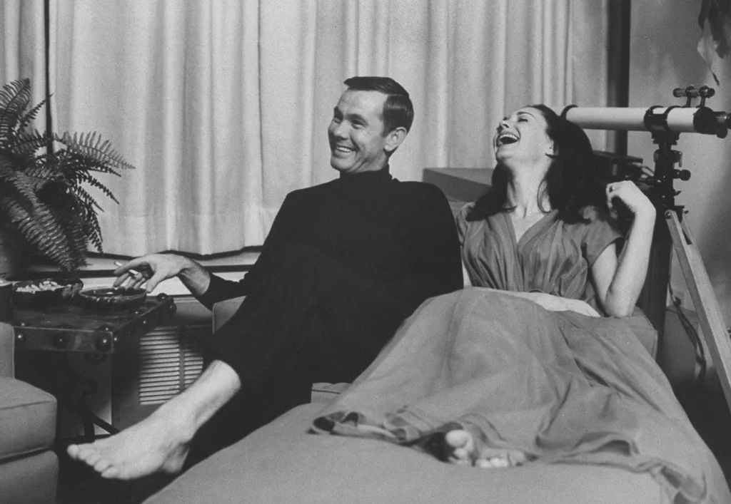 blackandwhite image of Johnny Carson legs crossed barefoot smoking seated beside his laughing wife in living area of...