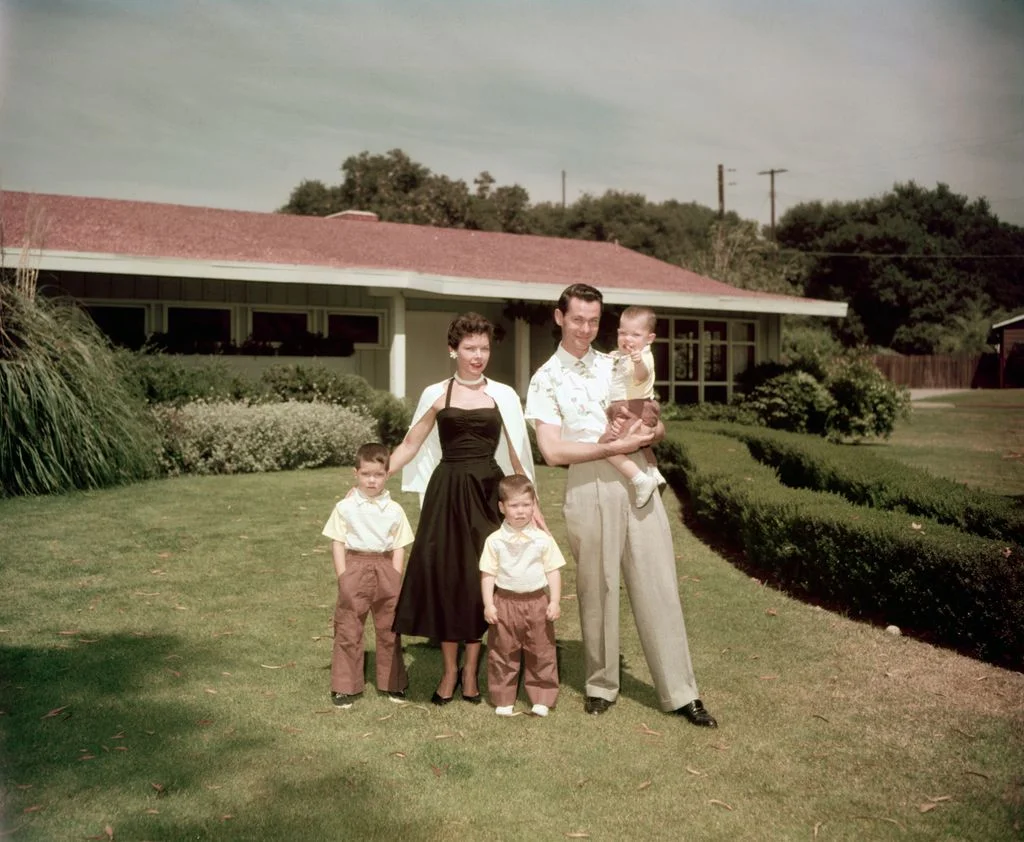 Johnny Carson with wife Jody Wolcott and three young songs standing on front lawn of ranchstyle home