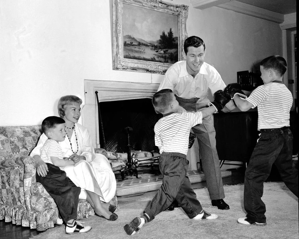blackandwhite photo of Johnny Carson play fighting with his two eldest sons in living room the youngest sits to left...