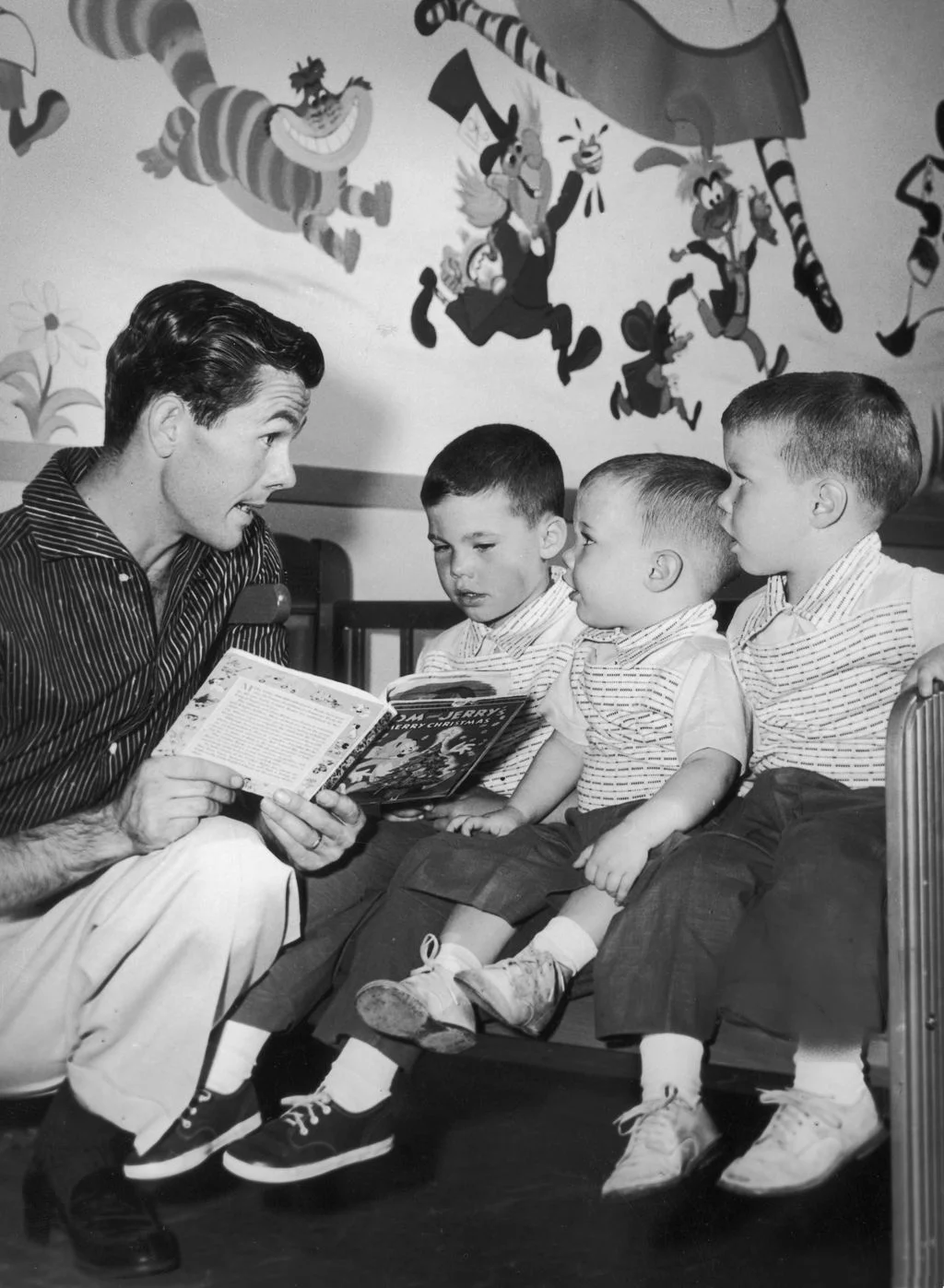 blackandwhite photo of Johnny Carson left knelt down reading to three young sons who sit on a bed facing him mural of...