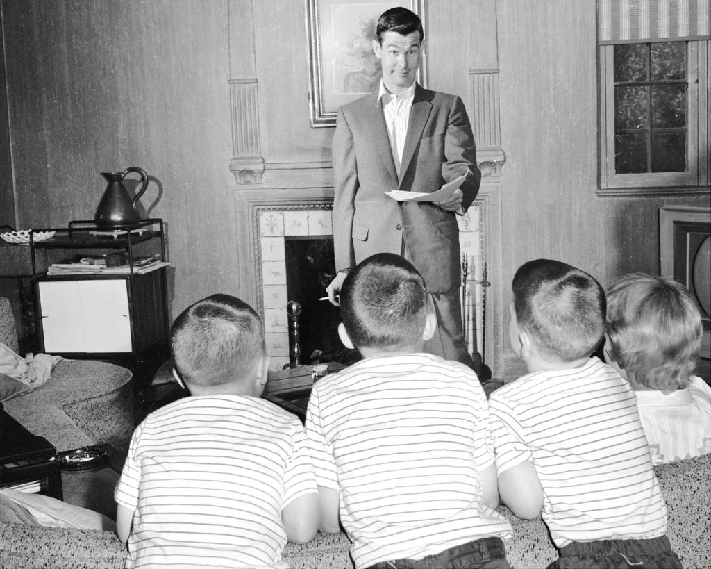 blackandwhite photo of Johnny Carson stood up reading off a script to his three sons wearing matching striped shirts...