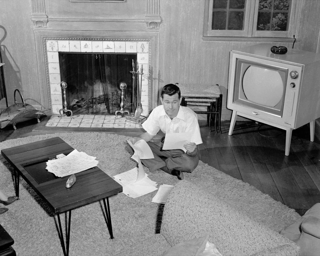 blackandwhite photo of Johnny Carson seated on living room rug going over papers hardwood floor TV to right fireplace...