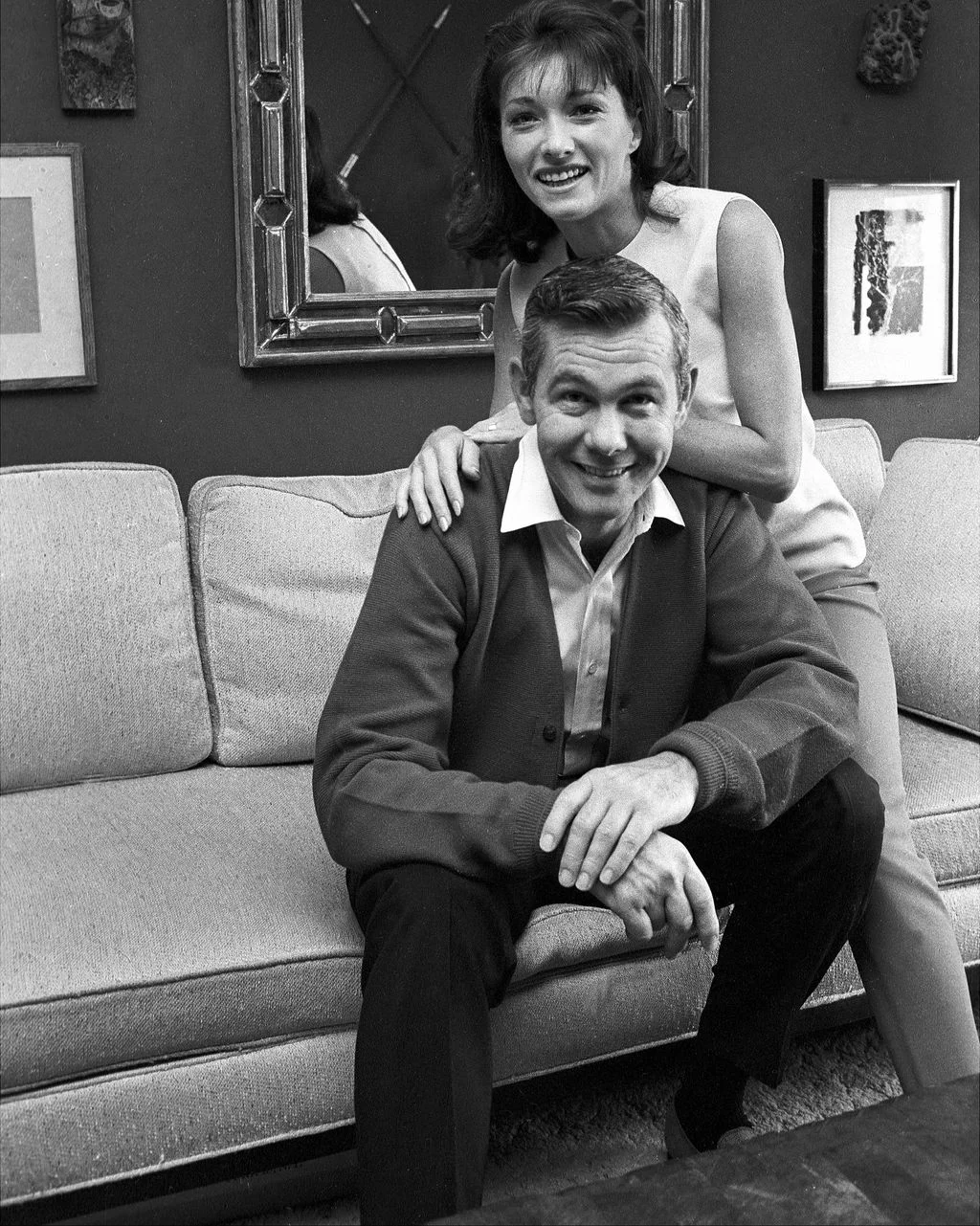 blackandwhite photo of Johnny Carson seated on sofa smiling wife Joanne rests her hands on his shoulders knelt down...