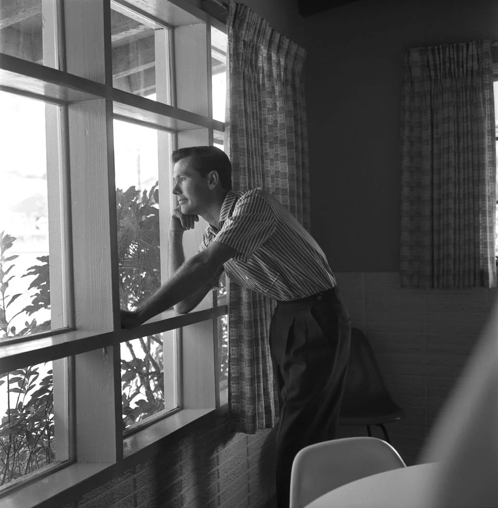blackandwhite photo of Johnny Carson looking out window of his home dining table to right