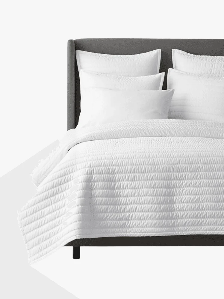 White sateen quilted coverlet on a bed with several pillows up against a gray headboard.