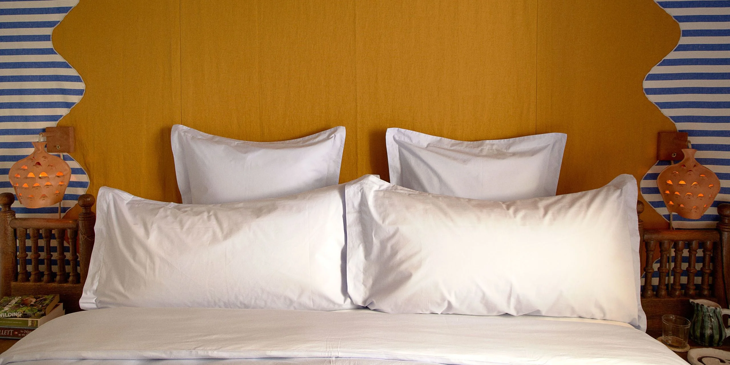 Head of a bed with white bedding in front of a yellow and striped wall.