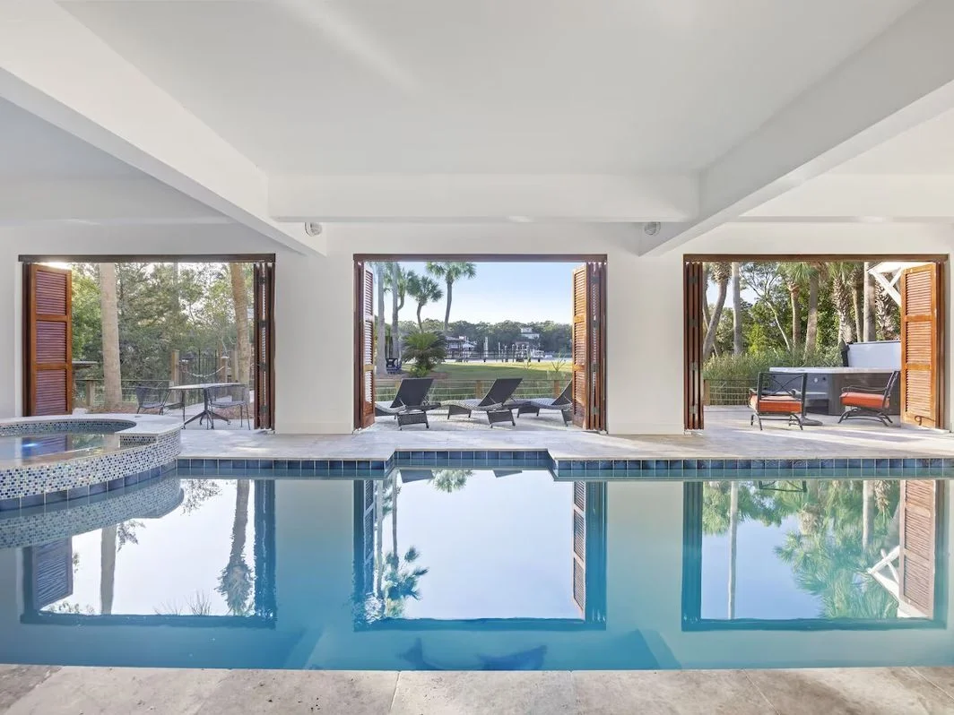 An indoor pool features a wall of open patio doors to the outside