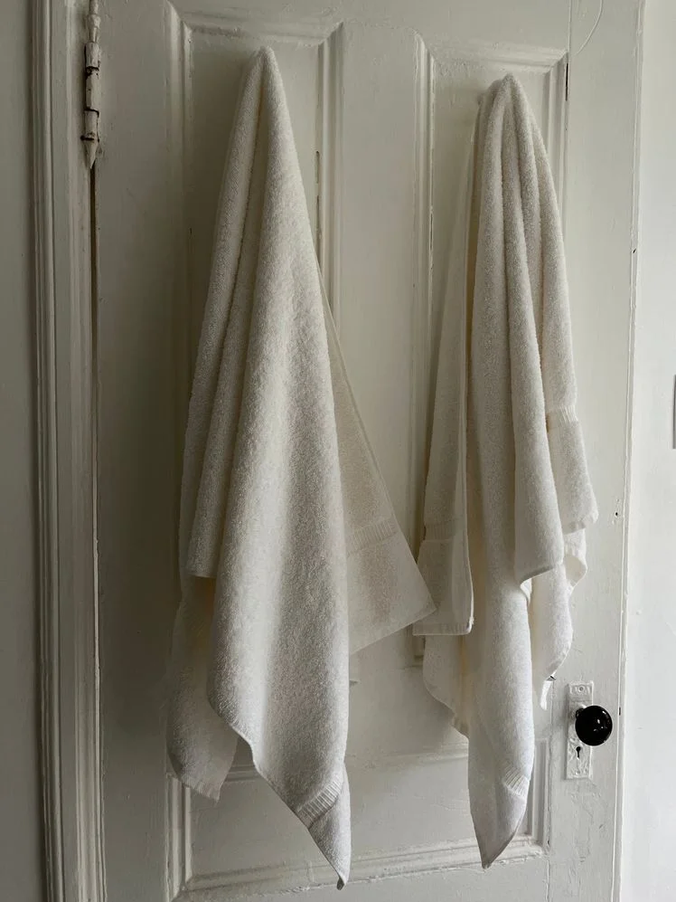 Two white bath towels hanging off hooks on the back of a bathroom door.