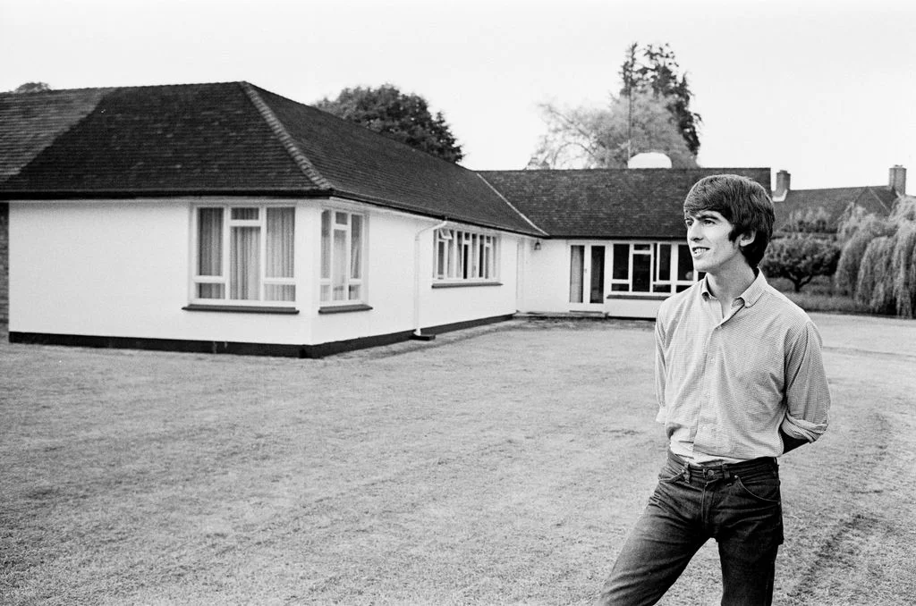 George Harrison smiling with arms behind back standing on lawn looking to left Lshaped home in background trees