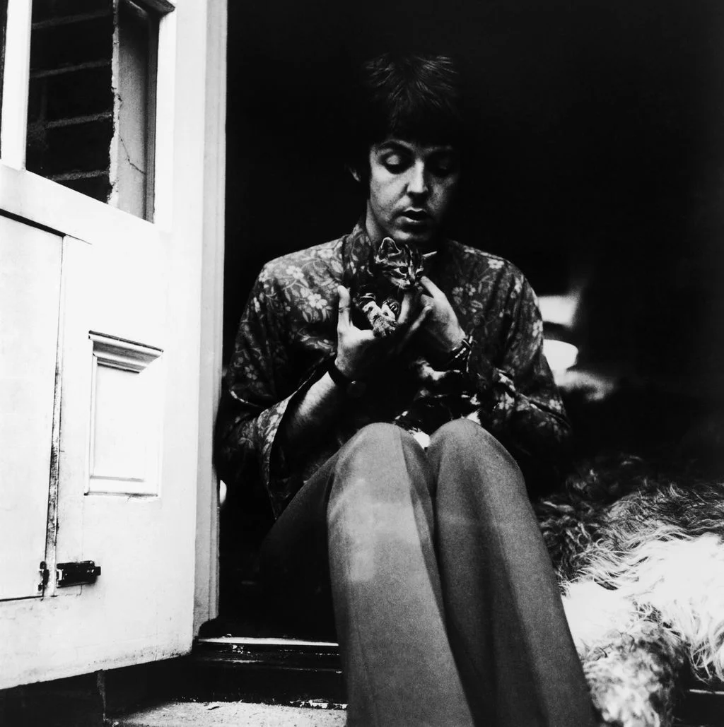 blackandwhite photo of Paul McCartney seated in doorframe cradling two small kittens dog seated beside him to right
