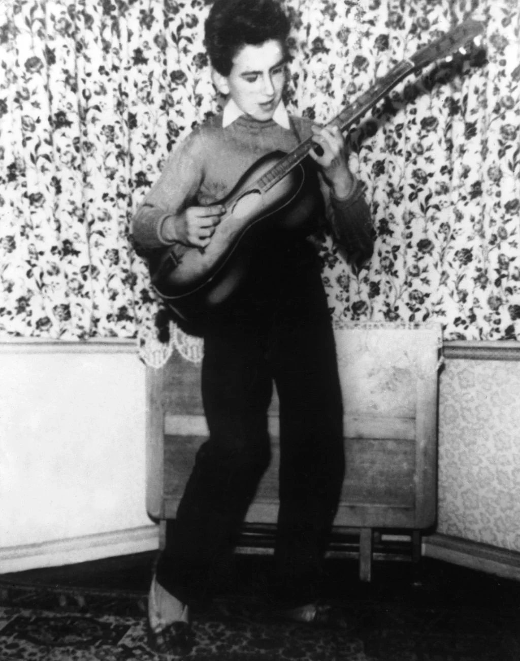 12yearold George Harrison  plays guitar standing in the corner of his house floral print wallpaper above wainscoting...