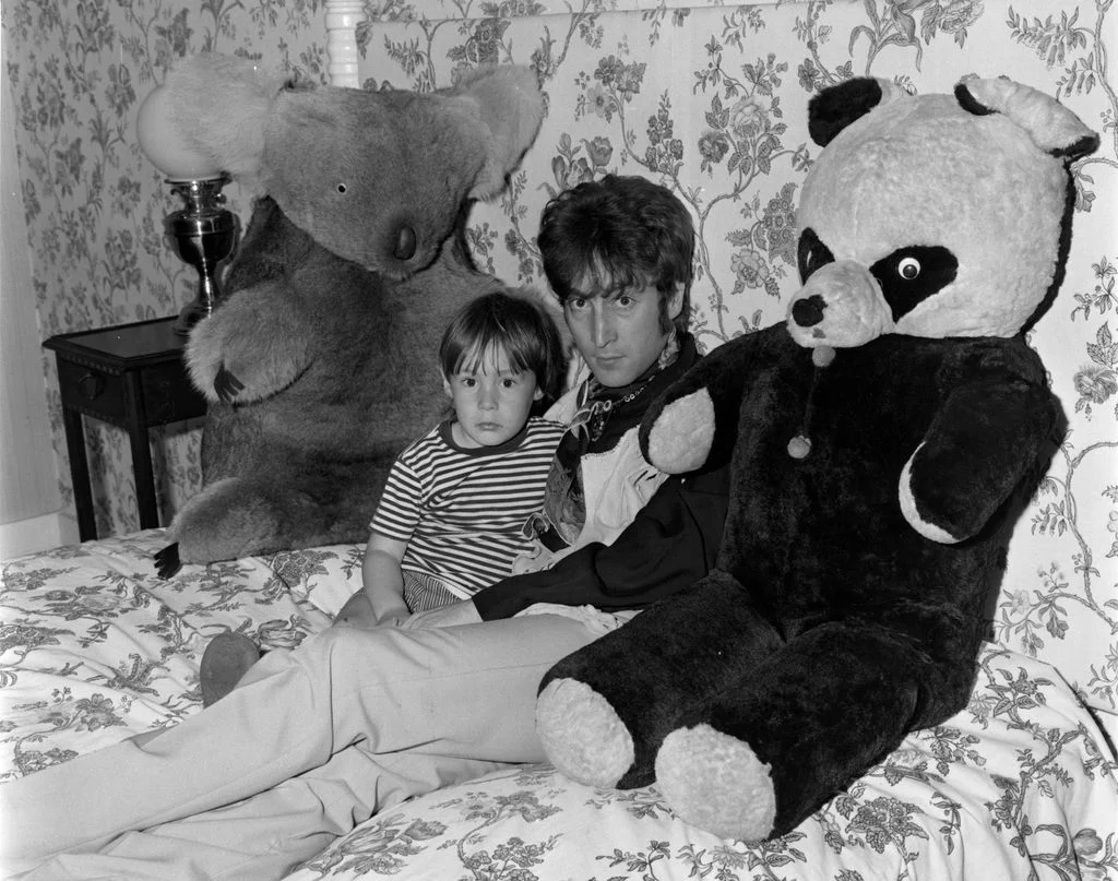 John Lennon sitting with young Julian Lennon between two large stuffed animals  on floral bedspread floral wallpaper...