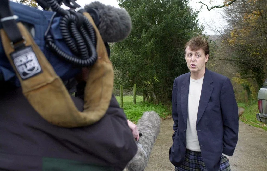 Paul McCartney stands with hands in pockets on dirt part lined with trees camera operator in foreground facing McCartney...