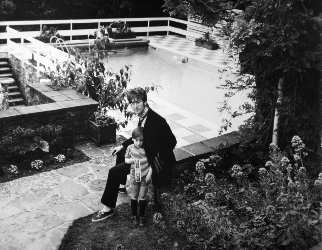 blackandwhite photo of John Lennon sitting on stone ledge with young Julian Lennon beside their pool in background...