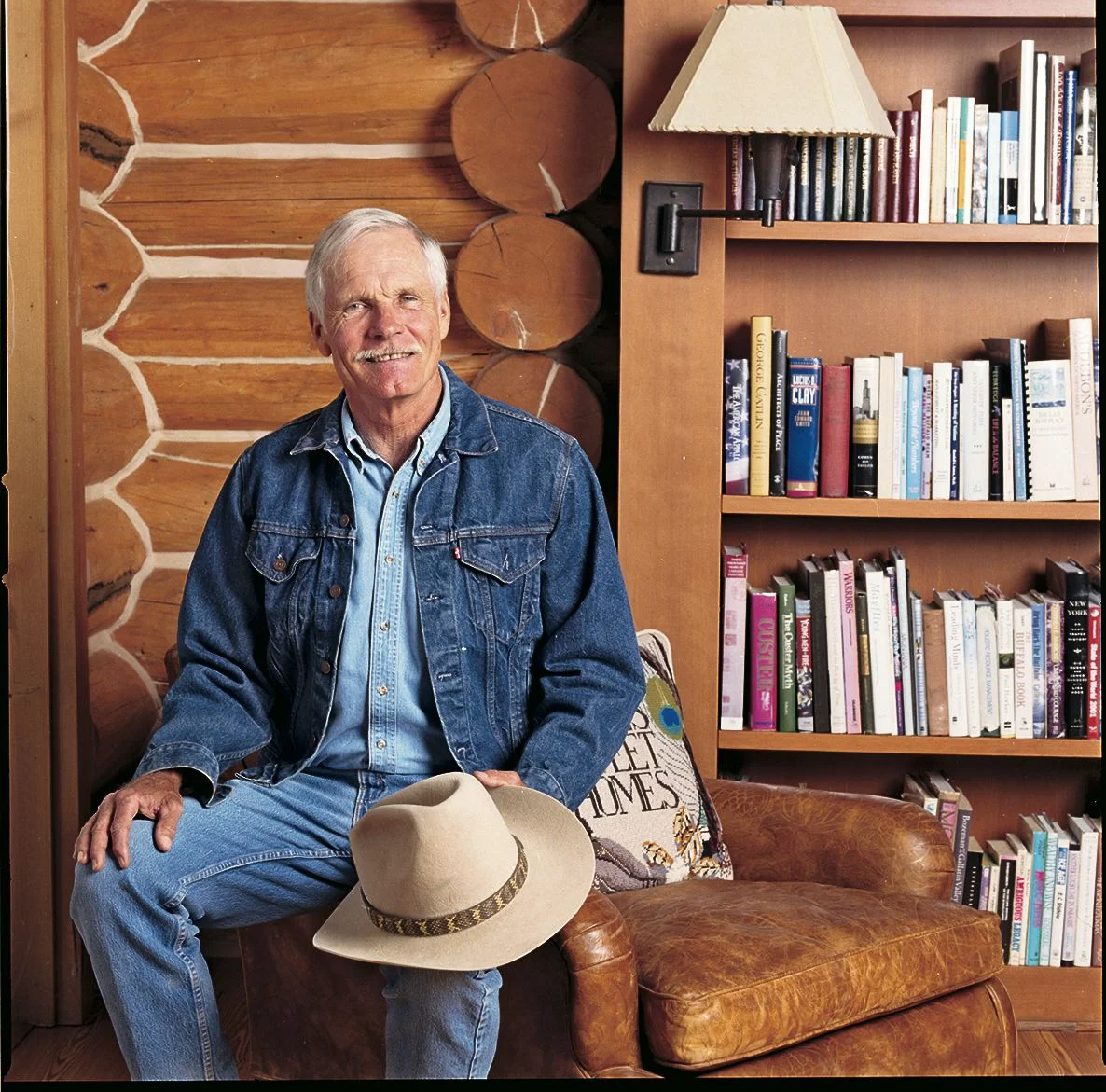 Image may contain Ted Turner Clothing Hat Adult Person Furniture Pants Sitting and Bookcase