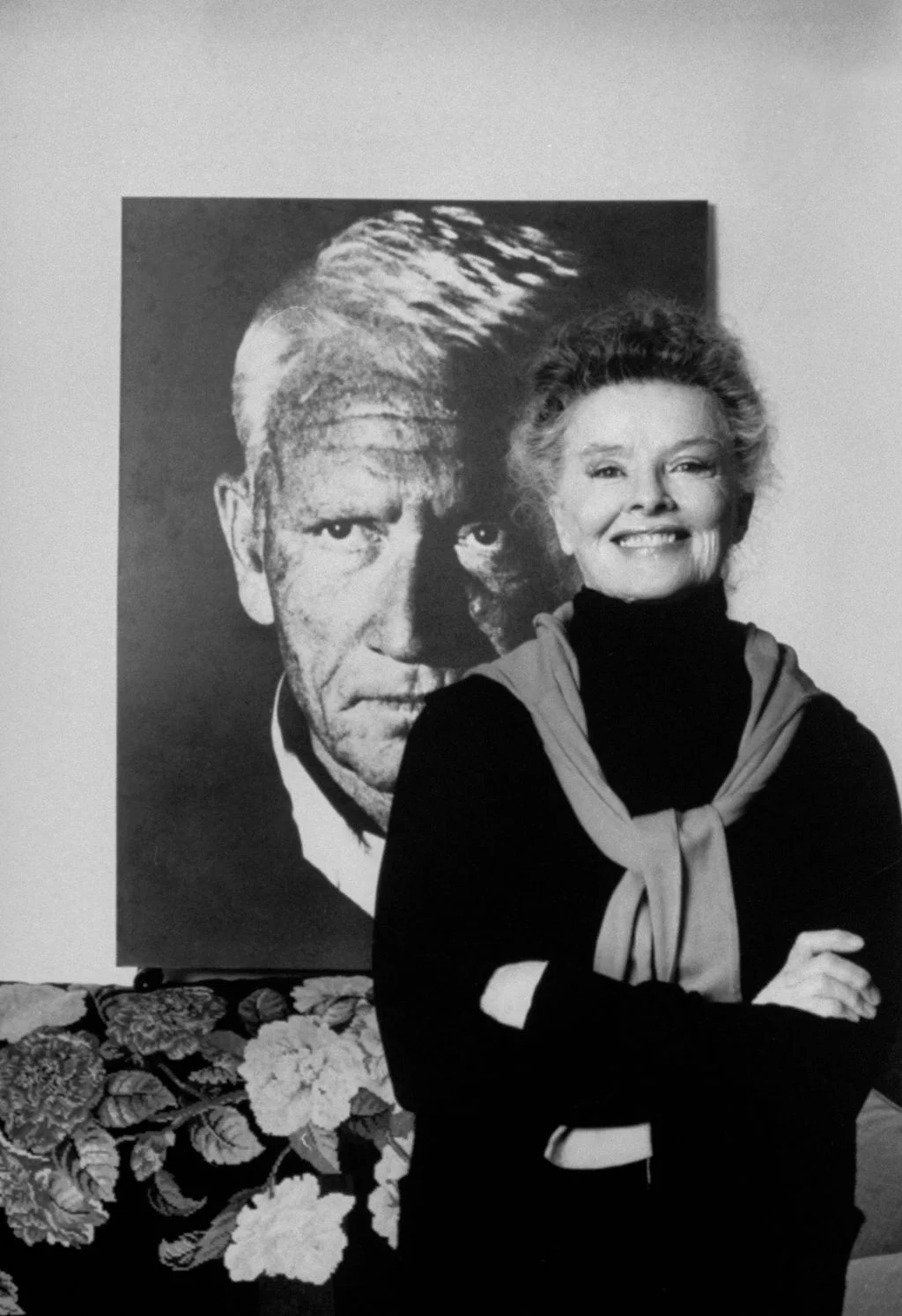 blackandwhite photo of Katharine Hepburn smiling arms crossed large photo of Spencer Tracy hanging in background