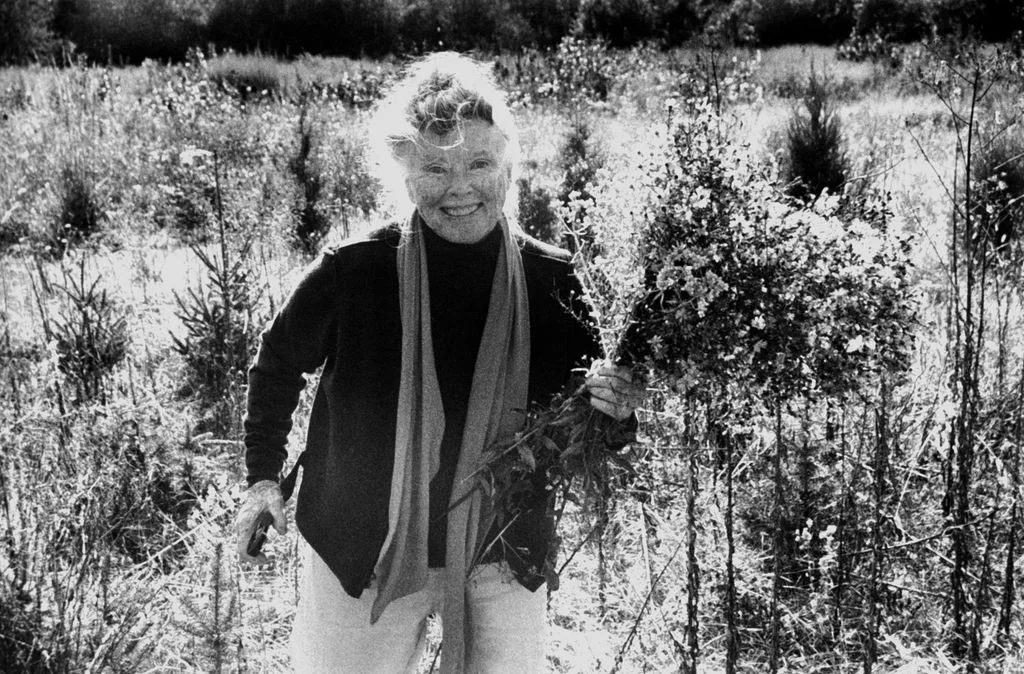 blackandwhite photo of Katharine Hepburn smiling standing in a field of wildflowers and greenery