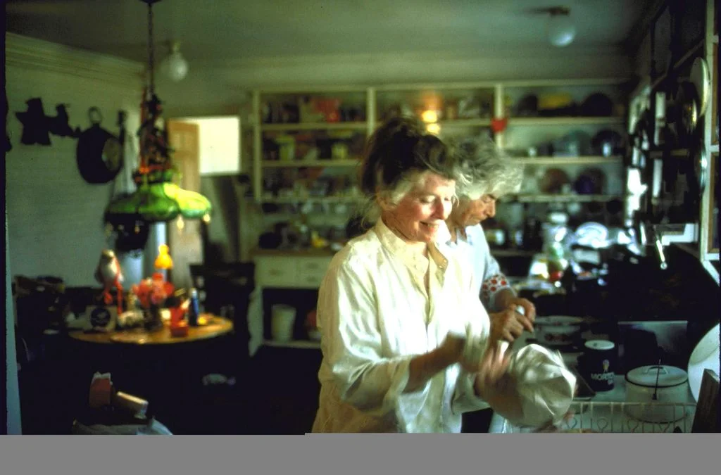 Katharine Hepburn in the kitchen drying dishes woman in background green lamp over round table in left background open...