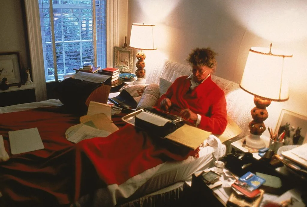 Katharine Hepburn in red sweater and blanket typing while seated on a typewriter at home
