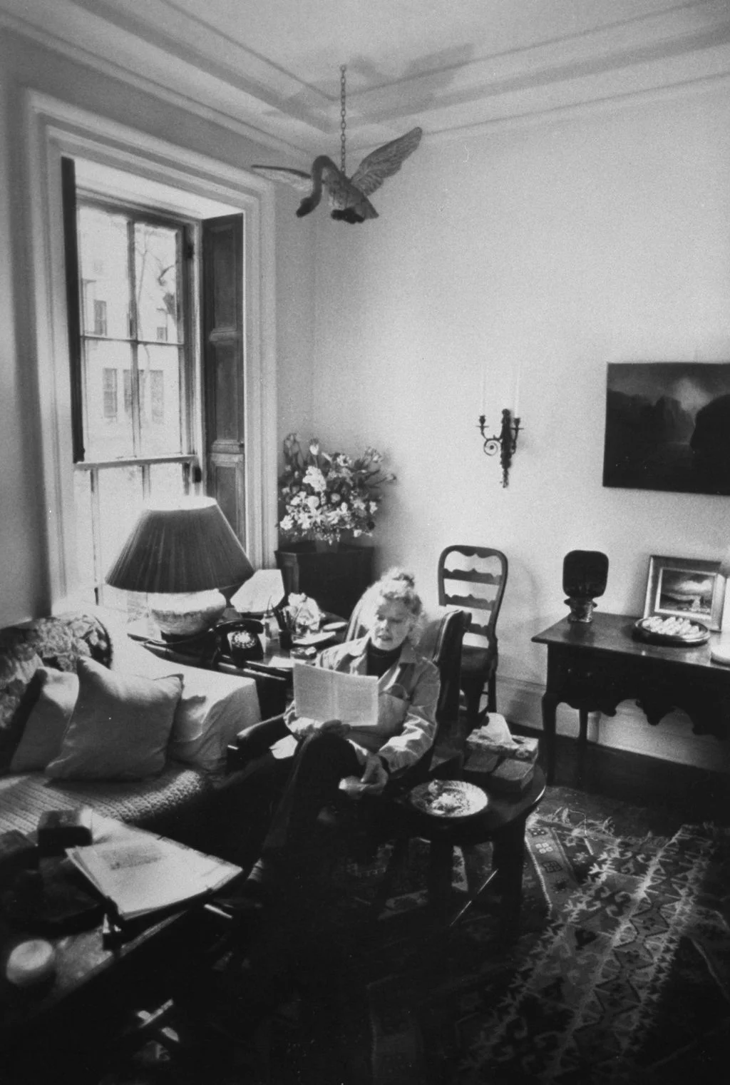 blackandwhite photo of Katharine Hepburn seated at chair reading in her NYC home goose figurine hanging from ceiling...
