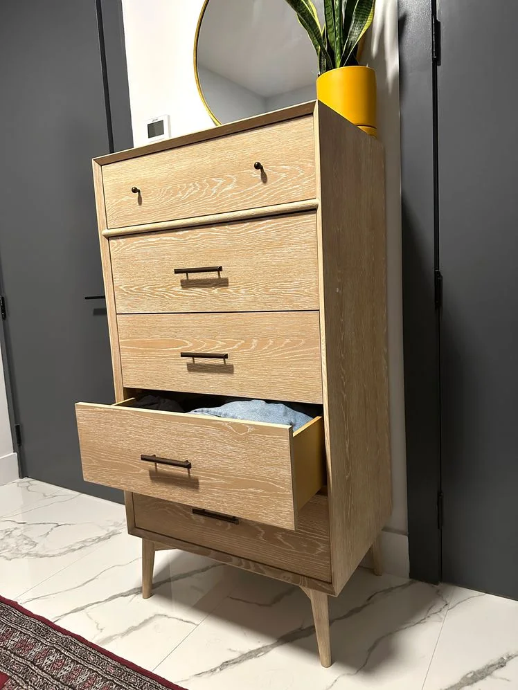 Tall pale wood dresser with one drawer open on marble floor in a bedroom.