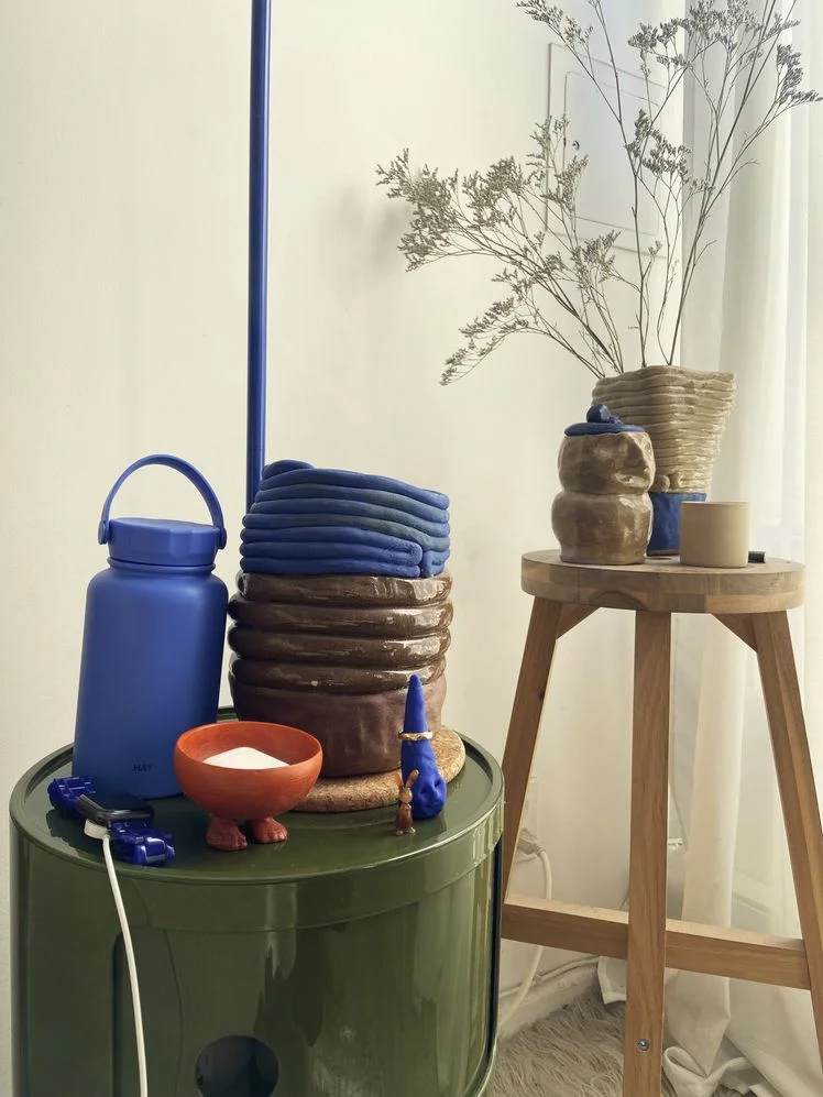 Cylindrical dark green nightstand next to a stool, both with coiled ceramic vases and bowls on top of them.