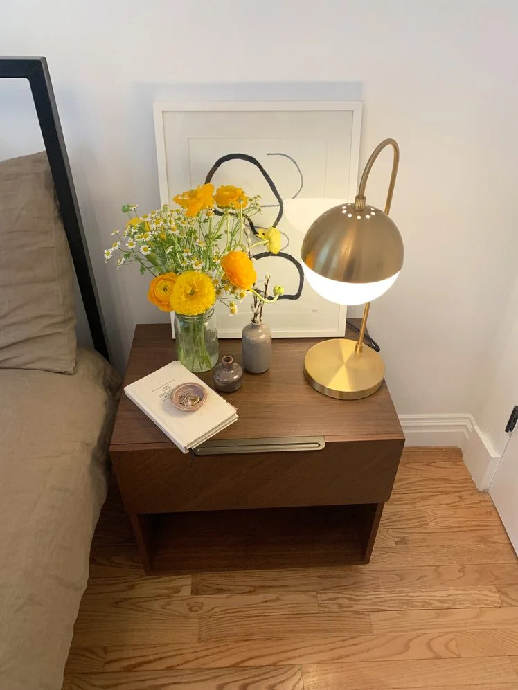 Nightstand next to a bed style with a vase of flowers, gold table lamp, white artwork, and two small ceramic vases.