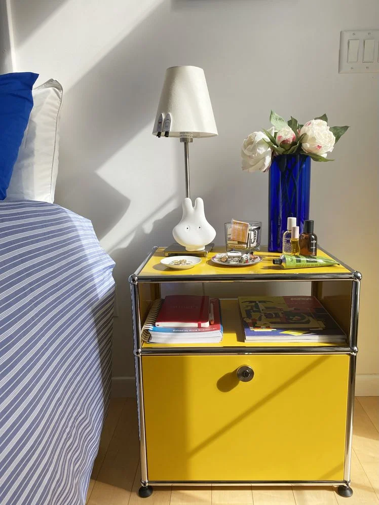 Yellow and silver nightstand styled with a lamp, vase of flowers, and assorted trinkets next to a bed made with blue bedding.